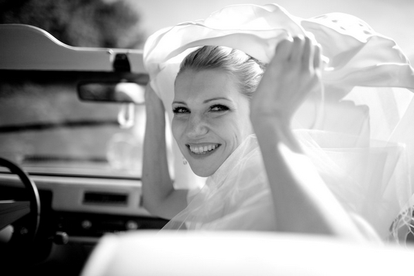 Bride in car holding veil as it blows in the wind - Picture by Wedding Photographer Duo
